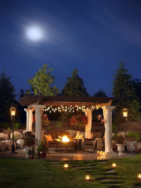 Pergola with string lights, fire pit, and cozy seating under a moonlit sky.