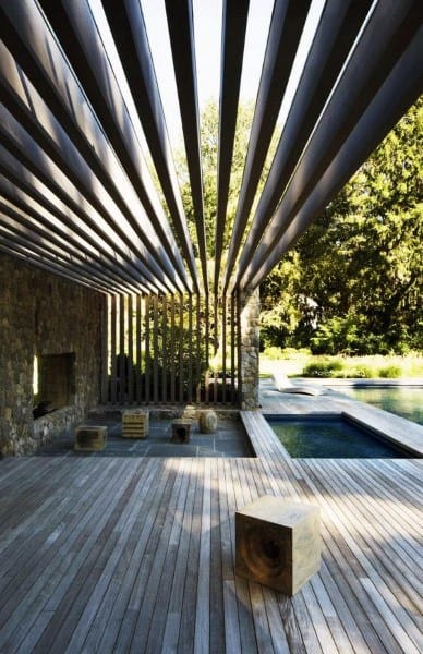 Modern pergola with bold slatted roof, stone fireplace, wooden deck, and poolside seating area.