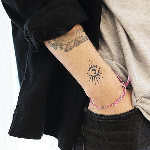 Minimalist tattoo of an all-seeing eye with radiating lines and a crescent moon, inked in fine black lines on the wrist.