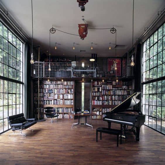 Spacious room with a grand piano, library shelves, and large windows; a mezzanine overlooks the wooden floor and seating area