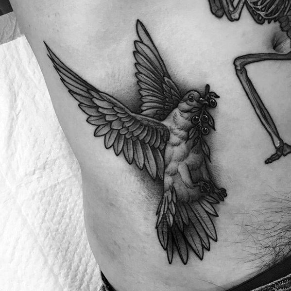 Black and grey pigeon tattoo in mid-flight holding an olive branch, symbolizing peace, hope, and strength, with bold linework and shading.