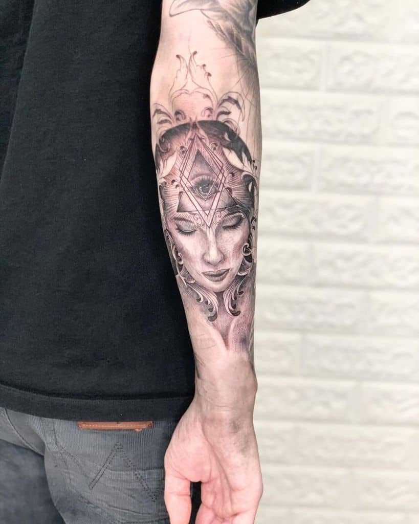 Tattoo of a mystical woman with a third eye inside a geometric design, surrounded by ethereal details, inked in black and grey realism on the forearm.