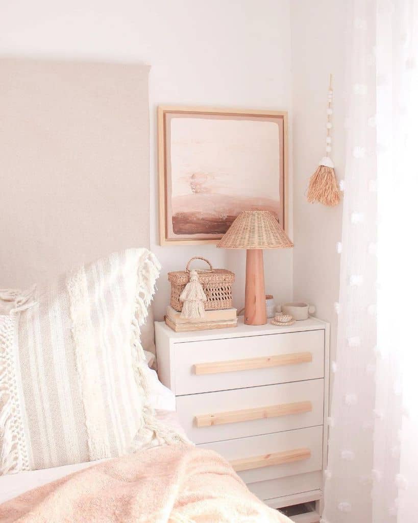 Cozy bedroom corner with a pastel-colored bed, a lamp, wall art, and a white dresser under soft, natural light