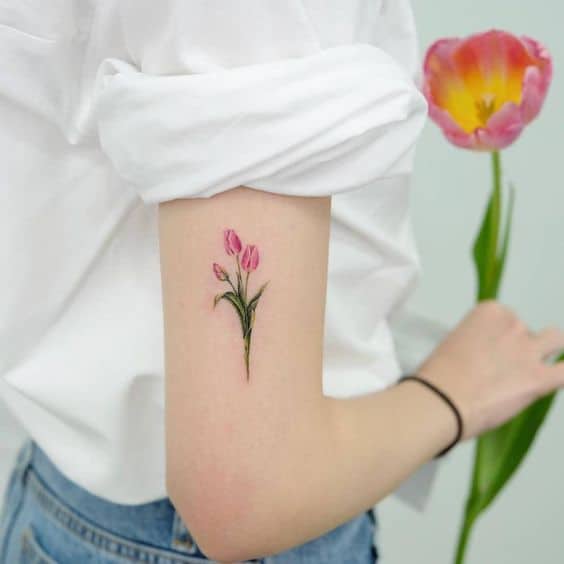 A delicate pink tulip tattoo on the upper arm, featuring two blooming flowers with fine details and soft green leaves in a realistic style.