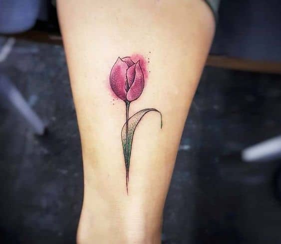A watercolor-style tulip tattoo on the thigh, featuring a vibrant pink flower with soft shading, a curved green stem, and artistic ink splashes.