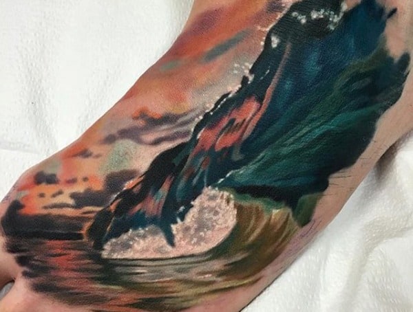 Foot tattoo for men featuring a colorful ocean wave crashing under a vivid sunset sky in a realistic painting style.