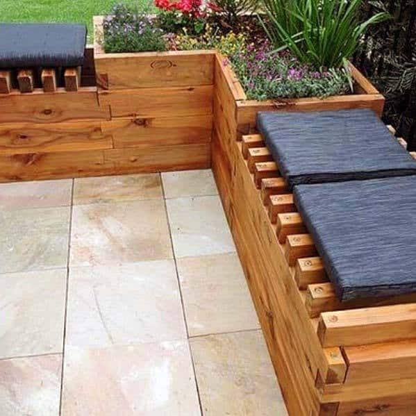 A wooden bench with black cushions, nestled among planters filled with flowers and greenery, graces the tiled patio