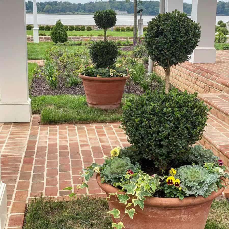 Potted topiary plants with flowers on a brick patio near a garden and water view in the background