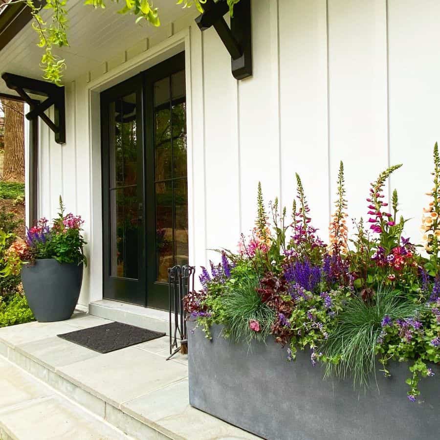A house entrance with black double doors, white siding, and large planters filled with colorful flowers on the porch