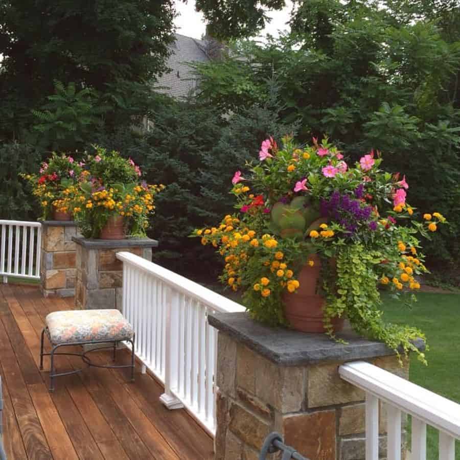 Wooden deck with two large flower pots filled with colorful blooms and greenery, bordered by a stone and white railing