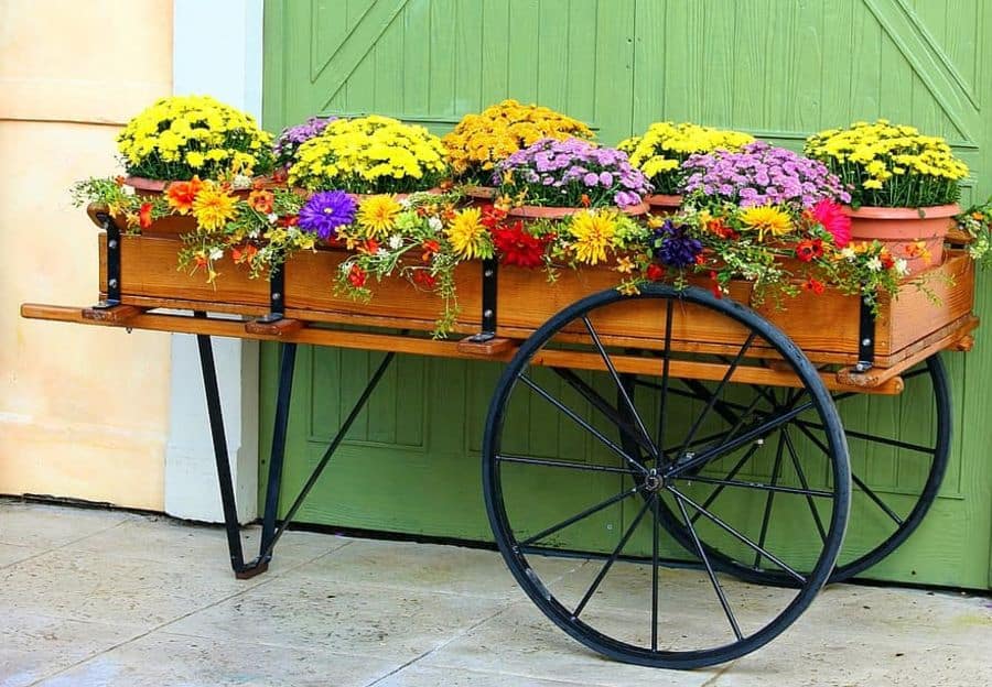 A wooden cart with colorful flowers in pots, featuring yellow, pink, purple, and red blooms, stands against a green door
