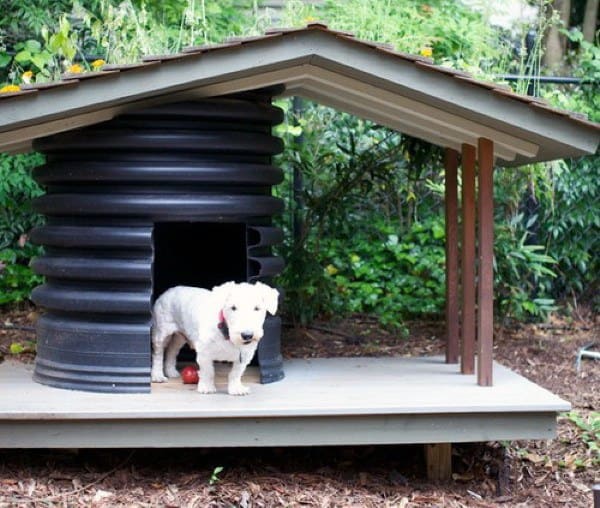 A unique dog house made from a large plastic barrel for the base with a slanted roof and wooden support beams, with a dog standing in front of the entrance