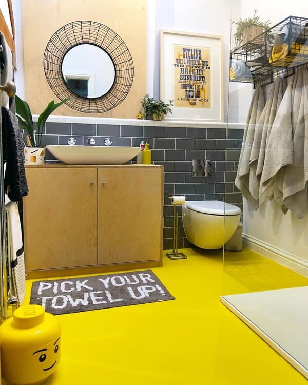 A bathroom with a vibrant yellow floor, plywood vanity, wire mirror, grey tile, and wire shelves storing towels.