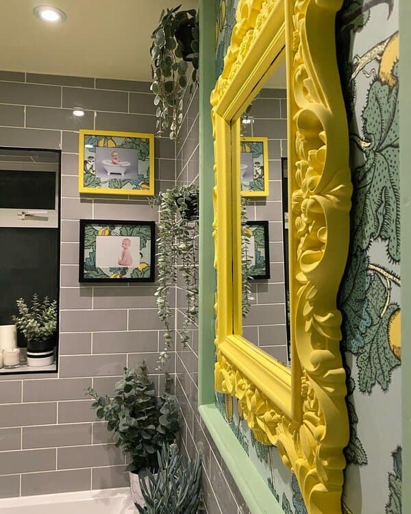 Bathroom with lemon-patterned wallpaper, a bright yellow carved mirror, grey subway tile, and hanging greenery.