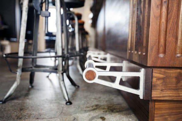 Polished steel foot rail with round design, mounted to a wooden bar with industrial-style metal stools on concrete flooring.
