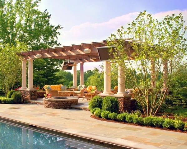 Poolside pergola with stone columns, cozy seating, and a firepit, surrounded by lush greenery.