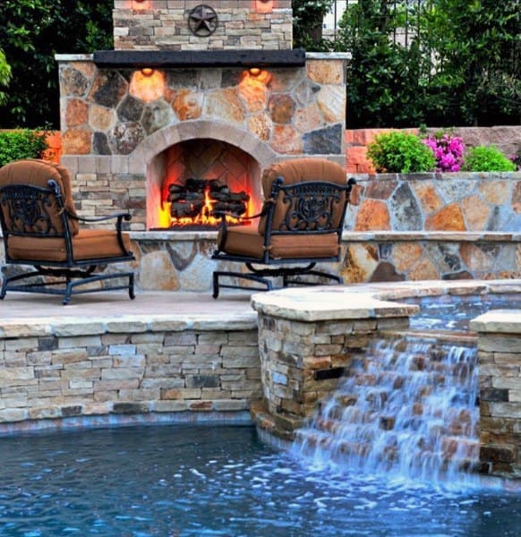 Outdoor stone fireplace with cozy chairs, cascading waterfall, and vibrant flowers nearby.
