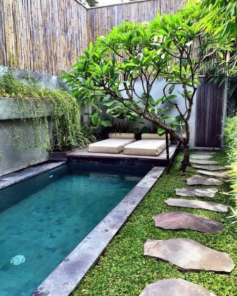 A serene rectangular pool with stone edges, a stone walkway, and a bamboo privacy fence in a lush garden.