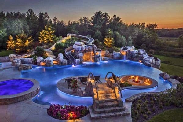 Luxurious outdoor pool with stone waterfalls, slide, fire pit, and surrounding greenery at sunset