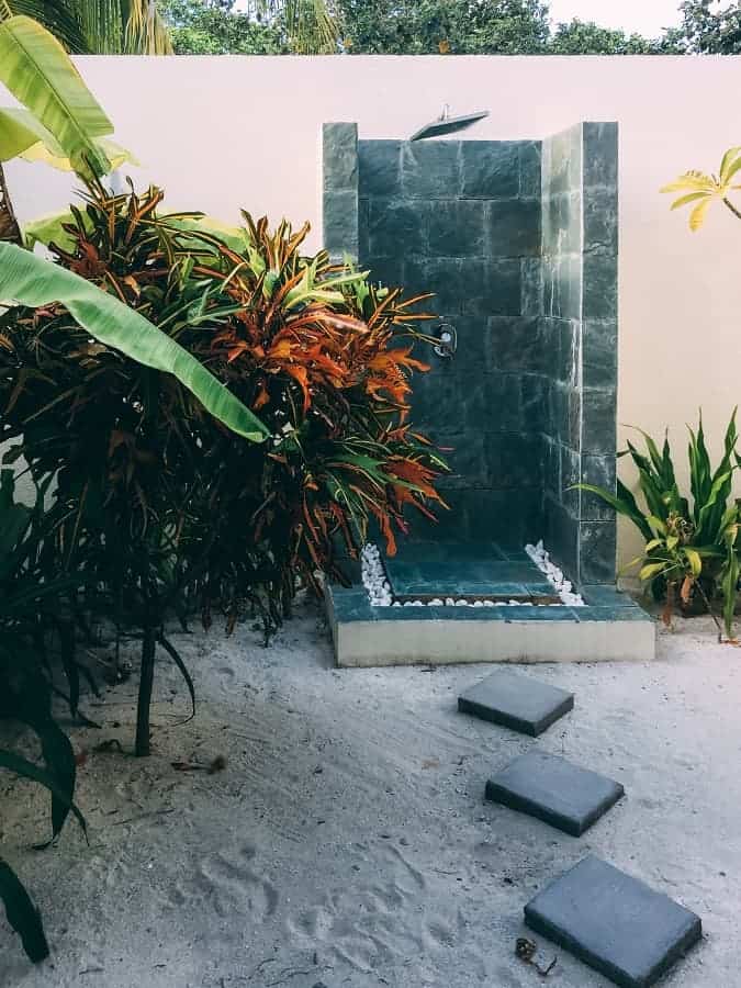 Outdoor shower with dark green tiles, surrounded by tropical plants and sandy stepping stones.