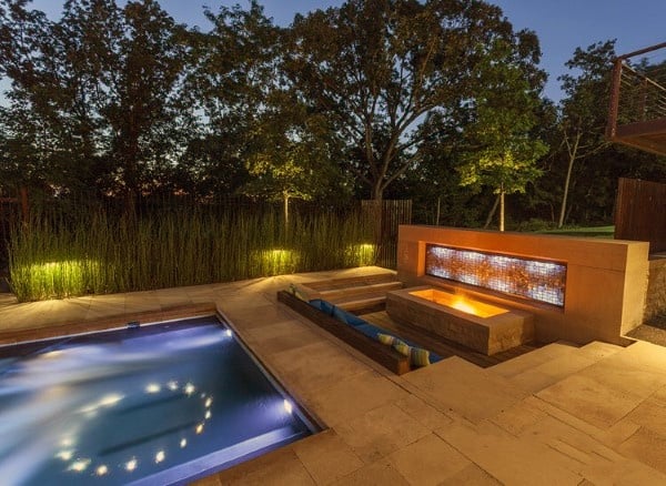 Sophisticated backyard design featuring a glowing gas fire pit, an illuminated pool, and lush landscaping.