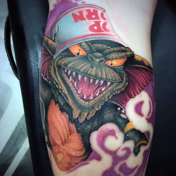 Gremlin tattoo with fierce details, glowing orange eyes, and a popcorn bucket hat, blending horror and humor in a vivid design.