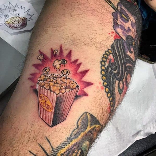 Tattoo of a bursting popcorn bucket with a red starburst background, flying kernels, and a bold POPCORN label on the container.