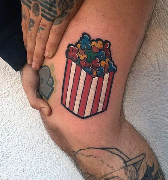 Tattoo of a red-striped popcorn bucket filled with colorful, rainbow-hued popcorn in a bold and playful cartoon style.