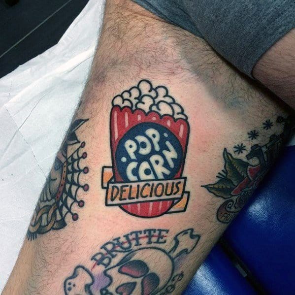 Tattoo of a vintage-style red popcorn bucket with the words POPCORN and DELICIOUS in bold lettering, surrounded by classic tattoo elements.