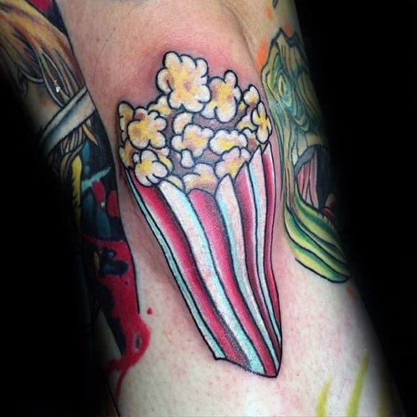 Tattoo of a tall red-striped popcorn bucket with fluffy yellow popcorn, done in a bold and vibrant cartoon style.