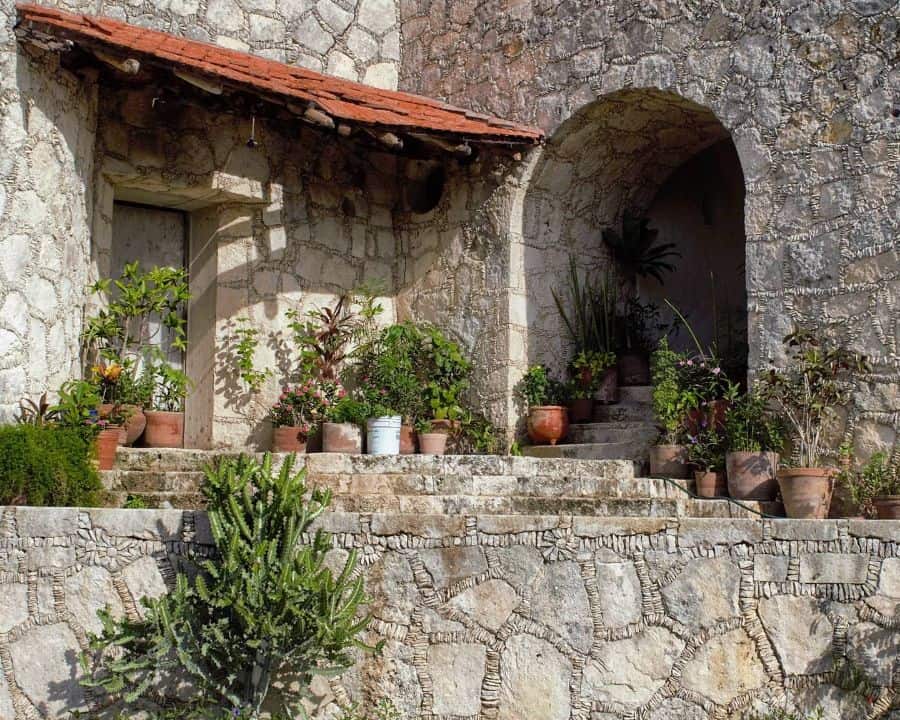 Stone building with an arched entrance, terracotta roof, and various potted plants on steps and ledges