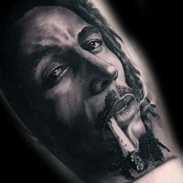 Hyper-realistic black and gray tattoo of Bob Marley smoking, with detailed facial shading, lifelike eyes, and smoke rising from the joint.