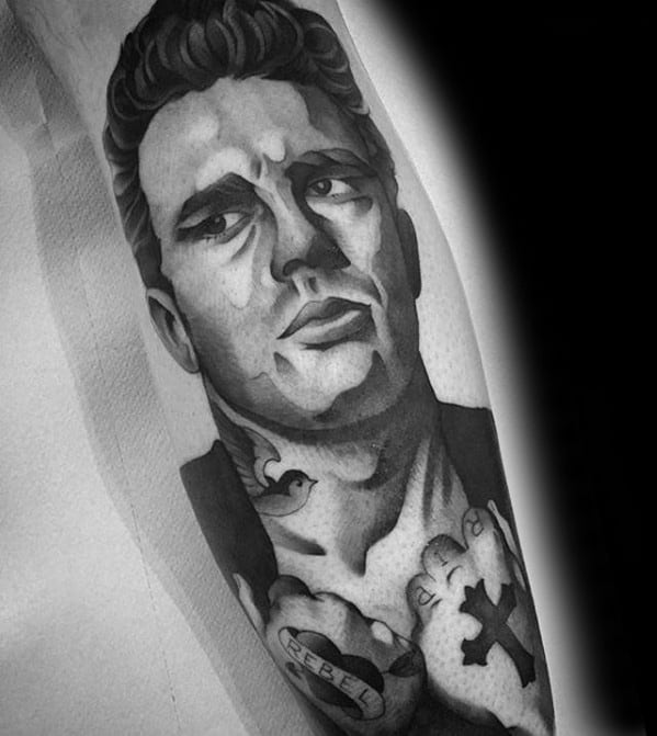 Black and grey neo-traditional tattoo of James Dean with bold shading, modern tattoos on his neck and fingers, and a cross and "REBEL" knuckle detail.