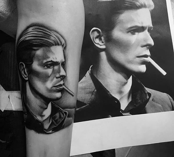Detailed tattoo of David Bowie in a contemplative pose, holding a cigarette, displayed alongside the original reference photo