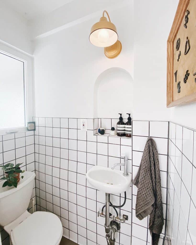 Small modern bathroom with white tiles, a mini sink, wall light, and a potted plant on toilet tank.