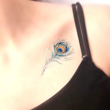 Small peacock feather tattoo on the collarbone, featuring delicate fine lines and soft blue, green, and gold hues for a subtle look.