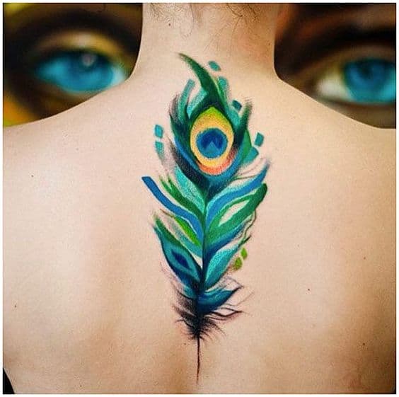 Watercolor-style peacock feather tattoo on the upper back, featuring vibrant blue, green, and yellow hues with soft brushstroke effects.