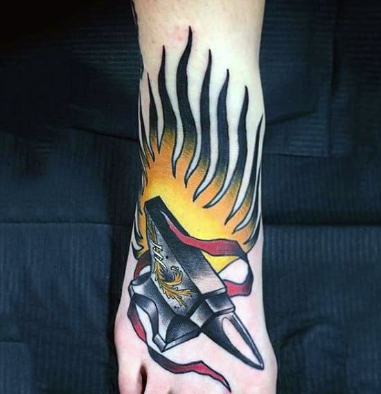 Tattoo featuring a stylized flame, an anvil, and ribbon on someone's foot