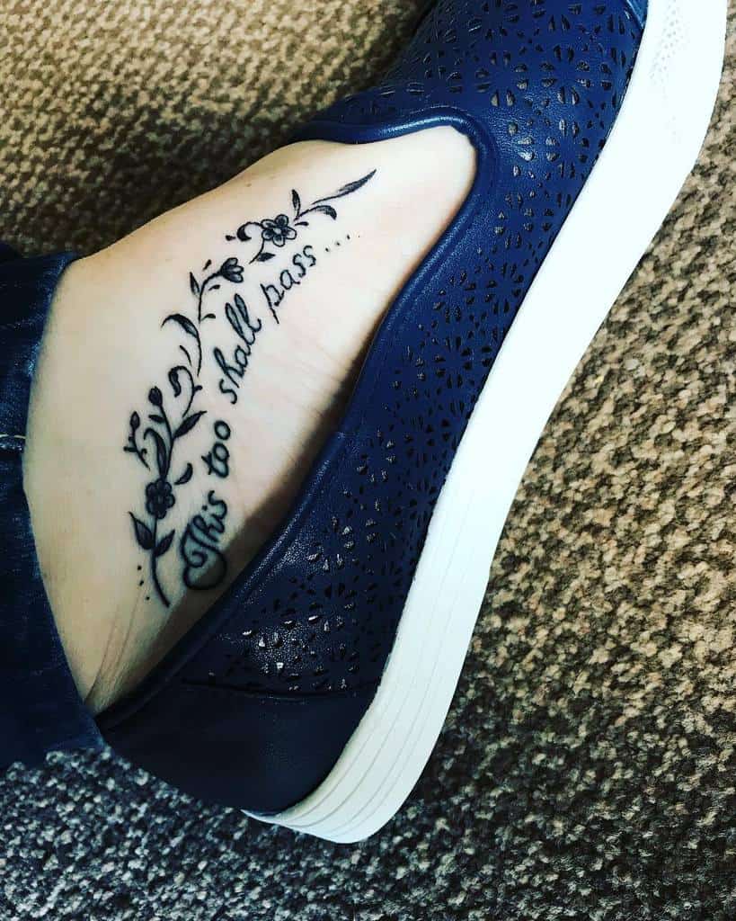 A "This Too Shall Pass" tattoo in cursive with flower designs, on the side of a person's foot.