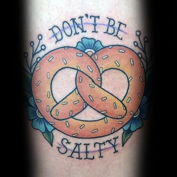 Golden pretzel tattoo with salt details, surrounded by blue flowers, featuring the phrase "Don't Be Salty" in bold lettering above and below.
