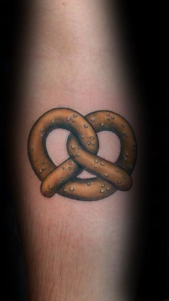 Realistic pretzel tattoo with rich brown shading and detailed salt specks, creating a three-dimensional effect on the forearm.