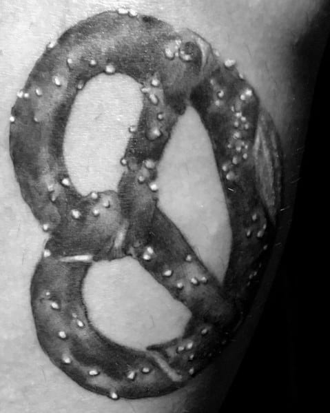 Black and grey realistic pretzel tattoo with detailed shading and salt specks, creating a textured and three-dimensional effect.