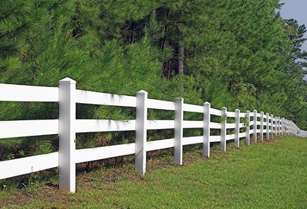 White farm-style post-and-rail fence bordering a grassy area with trees in the background.
