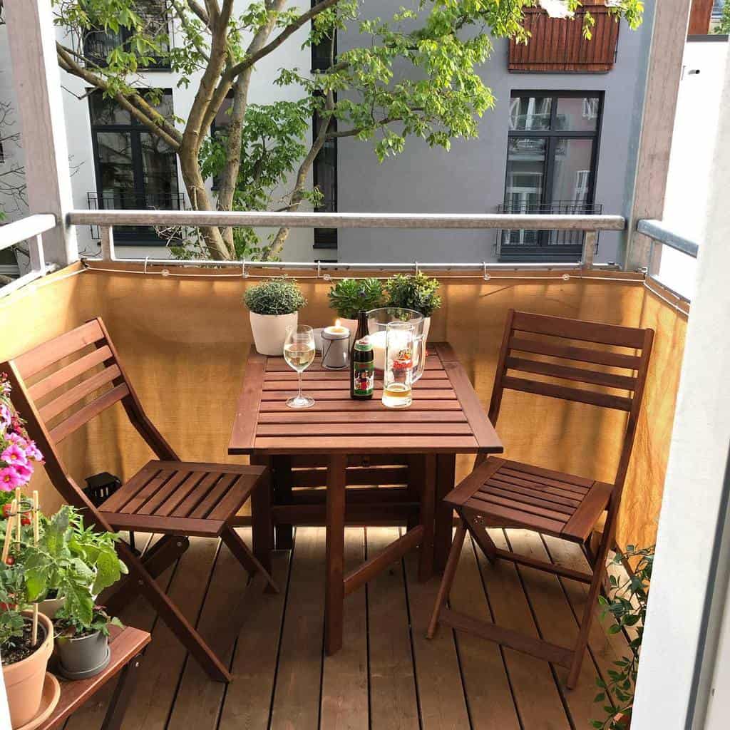 Small balcony with wooden table, chairs, and potted plants in a cozy urban setting.