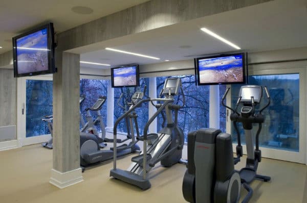 A modern gym with exercise machines, TV screens, and homey vibes, surrounded by windows showing a snowy landscape outside