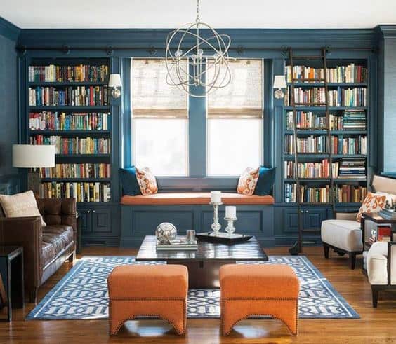 Cozy living room with blue walls, library shelves, window seat, orange ottomans, and patterned rug