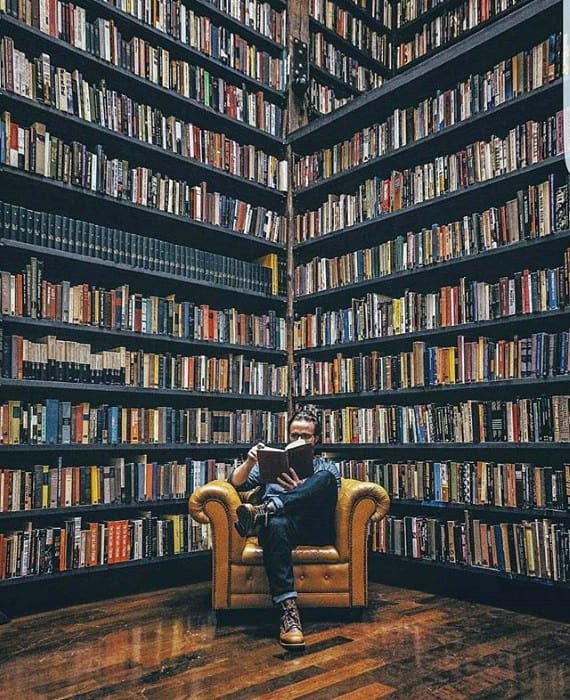 Man reading in a cozy armchair surrounded by towering bookshelves filled with books in a library