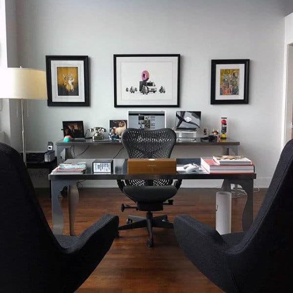 Modern home office with a glass desk, black chair, artwork on the walls, and various items on the desk