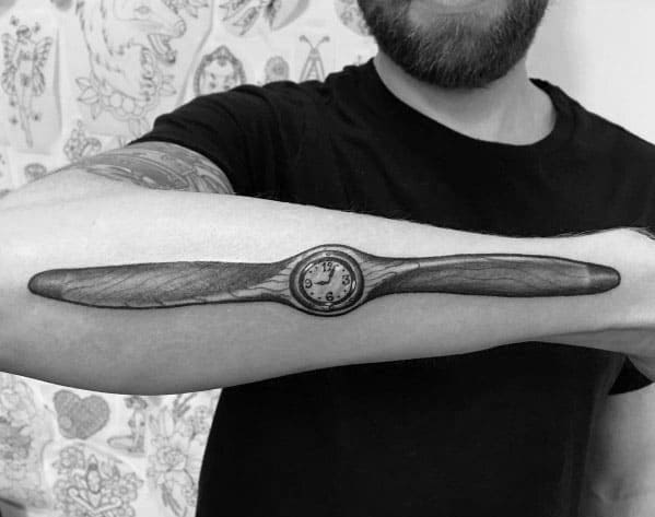 Realistic black and grey forearm tattoo of a vintage wooden propeller with intricate shading and a built-in clock at the center for a unique aviation theme.
