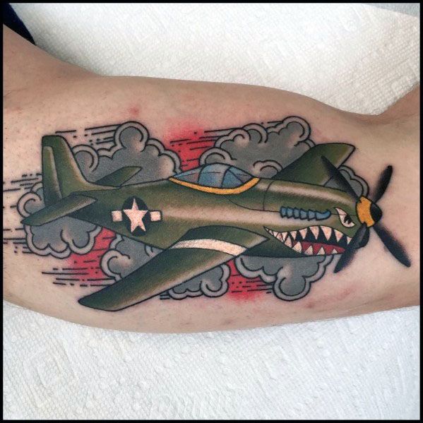 Colorful bicep tattoo of a WWII fighter plane with a shark mouth design, bold outlines, dynamic clouds, and red accents for a classic aviation look.